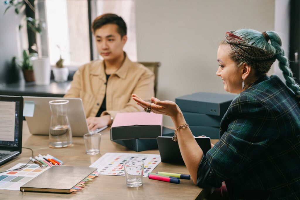 Two colleagues discussing a project at a desk with color swatches, markers, laptops, and stacked boxes in a bright office.